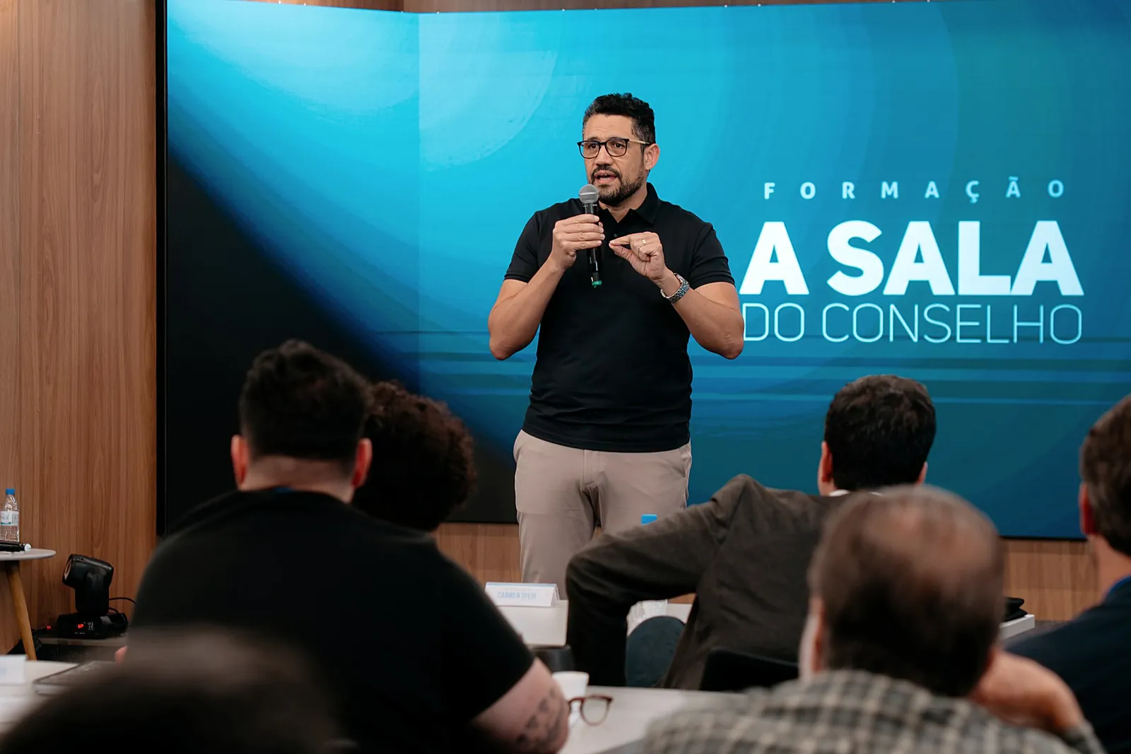 Vagner Campos dando aula diante do telão da Formação A Sala do Conselho, com a turma sentada à mesa