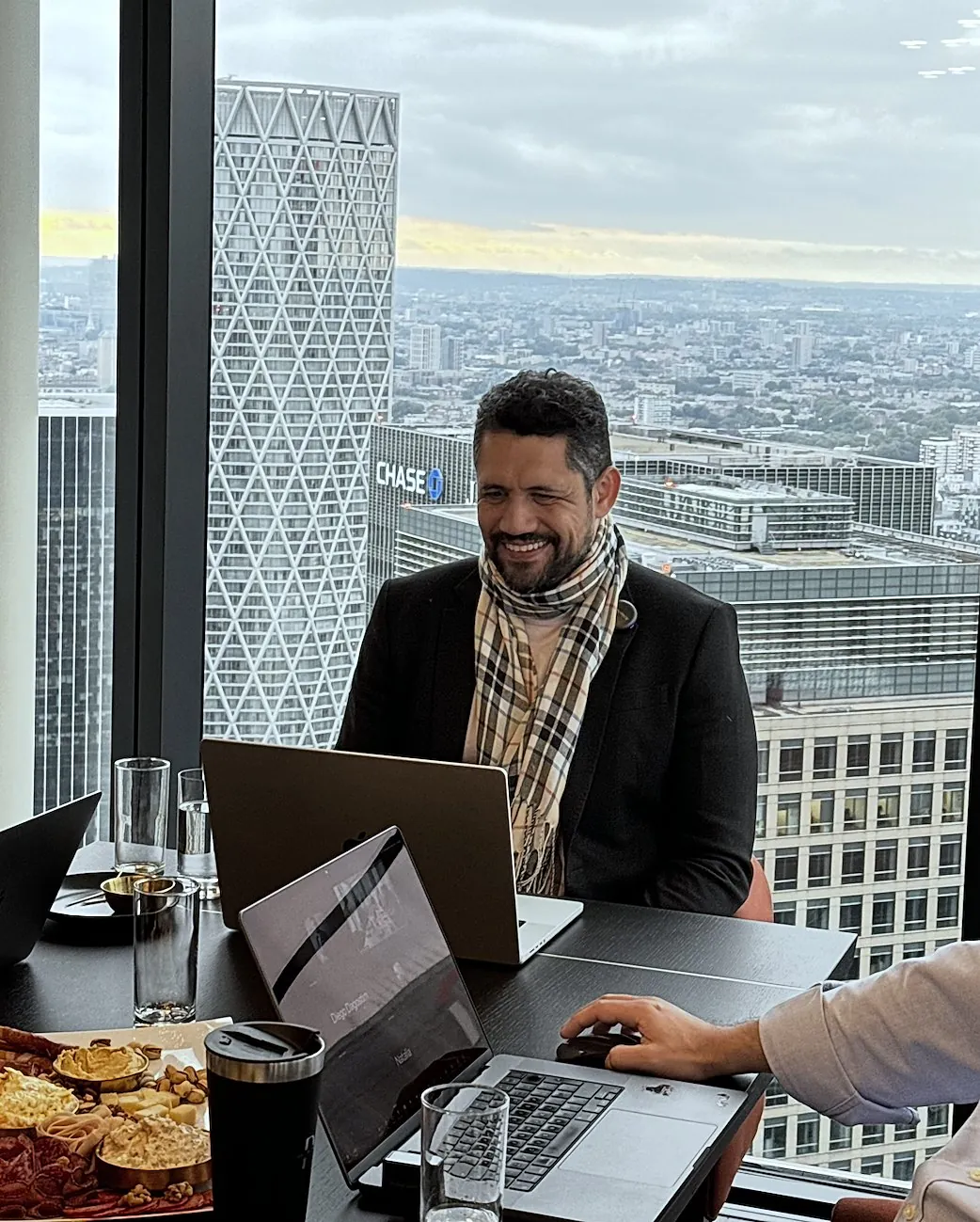 Vagner Campos sorrindo diante do notebook, com os prédios de Canary Wharf ao fundo
