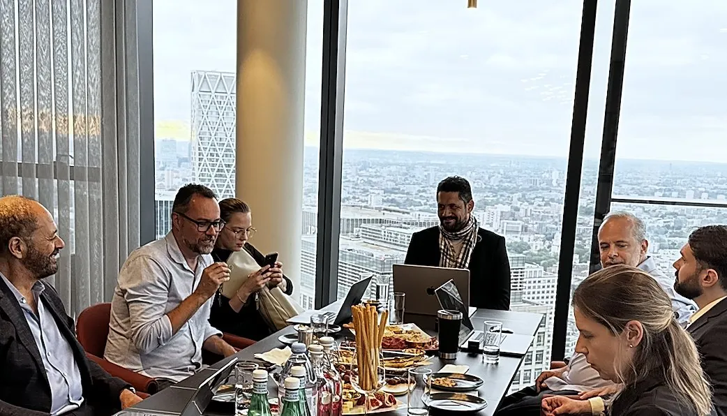 Empresários reunidos em torno de uma mesa em um andar alto de Canary Wharf, com Vagner Campos ao fundo diante do notebook