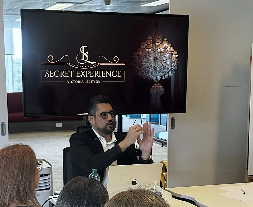 Vagner Campos explicando com as mãos diante de uma tela do Secret Experience, com participantes de costas